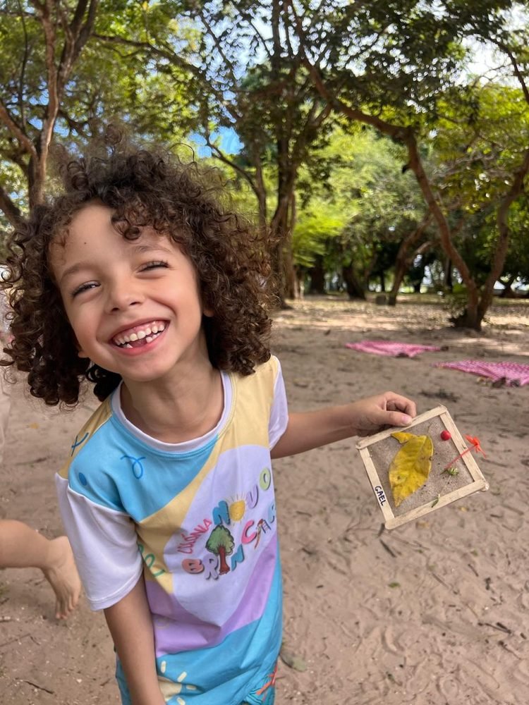 Criança sorridente com a camiseta do Novo Brincar, segurando um trabalho manual feito com folhas
